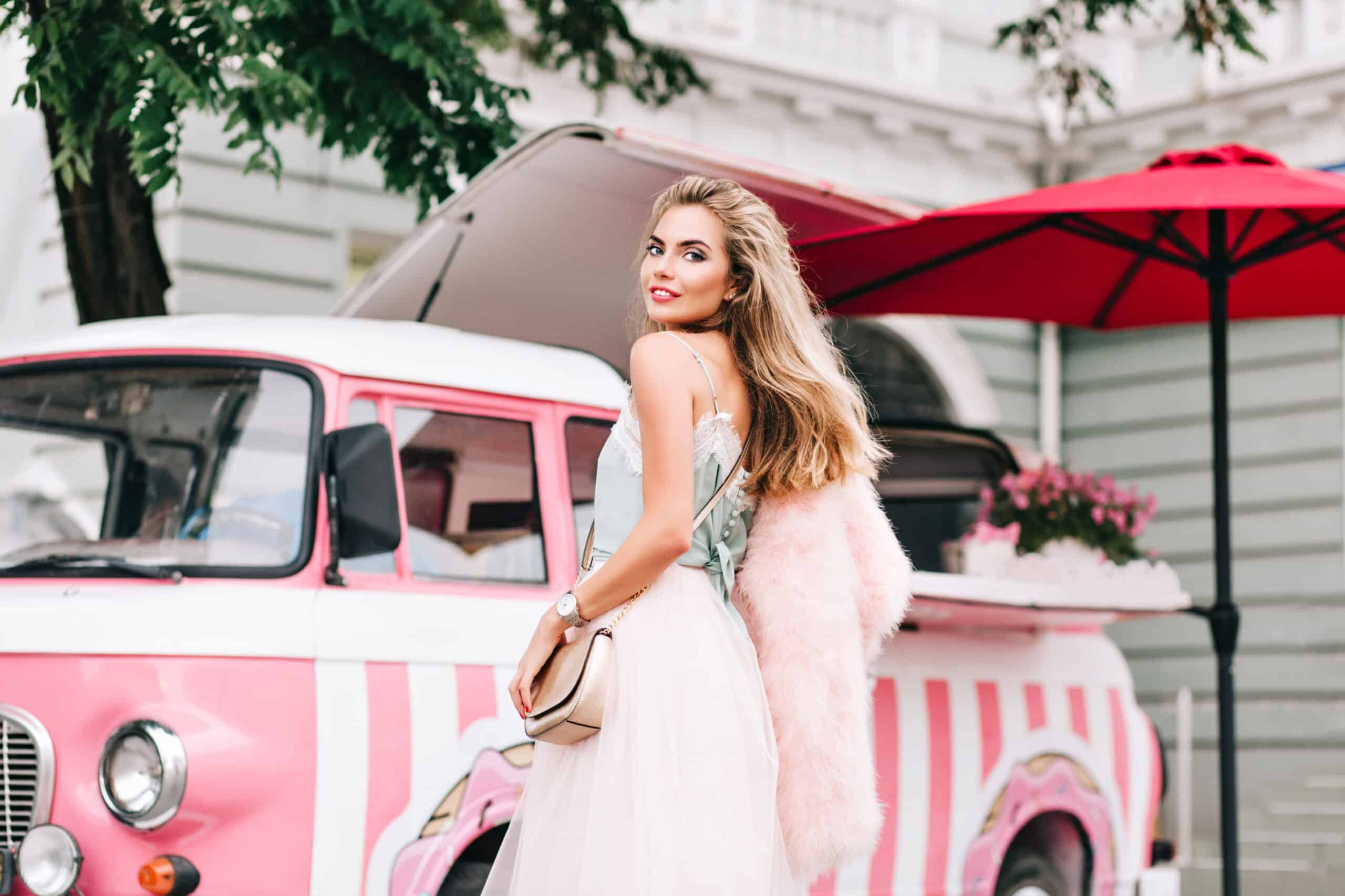 View from back fashion model in tulle skirt on retro coffee car background. She has long blonde hair, wears pink stole on shoulder, smiling to camera