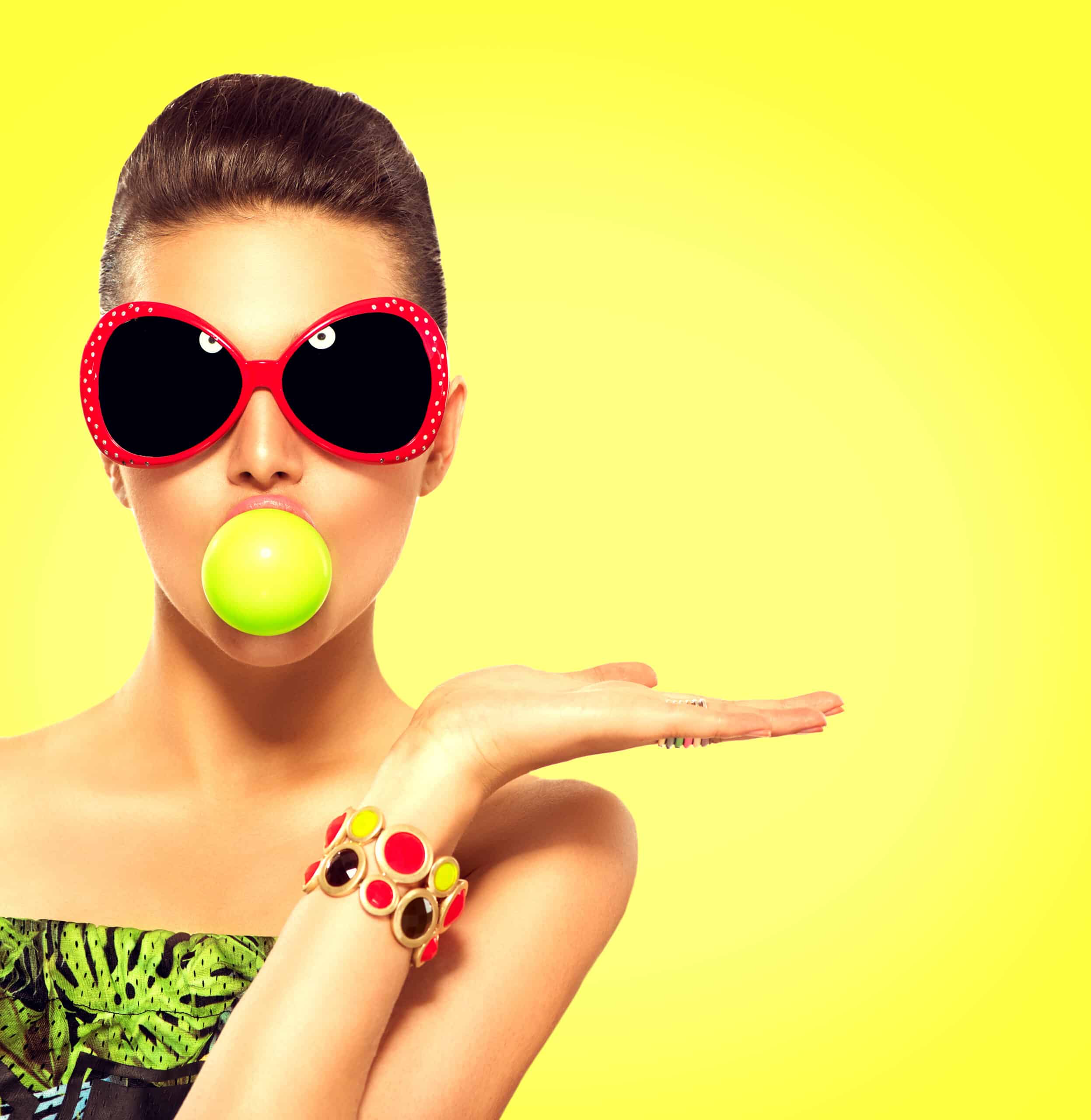 Beauty summer fashion model girl wearing sunglasses with green bubble of chewing gum and bright makeup showing empty copy space on the open hand palm for text over yellow background. Beautiful woman