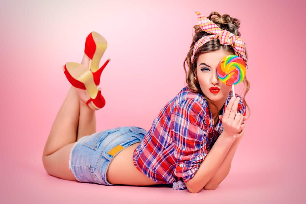 Sexy pin-up girl in shorts and high heels lying on a floor with bright lollipop over pink background. Beauty, fashion. Full length portrait.